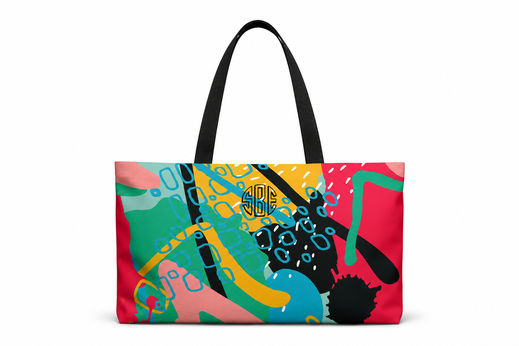 A tote bag with a vibrant abstract multicolor print featuring pink, teal, yellow, and black shapes, with monogram black shoulder straps, shown on a plain white background.