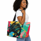 A woman wearing a white t-shirt and blue jeans carries a large tote bag with a colorful pop-art design featuring a Black woman's profile. The bag has a vibrant multicolor abstract background with pink, teal, yellow, and blue shapes, and black handles.