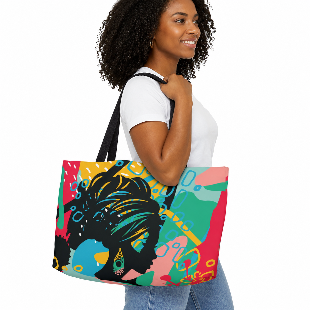 A woman wearing a white t-shirt and blue jeans carries a large tote bag with a colorful pop-art design featuring a Black woman's profile. The bag has a vibrant multicolor abstract background with pink, teal, yellow, and blue shapes, and black handles.